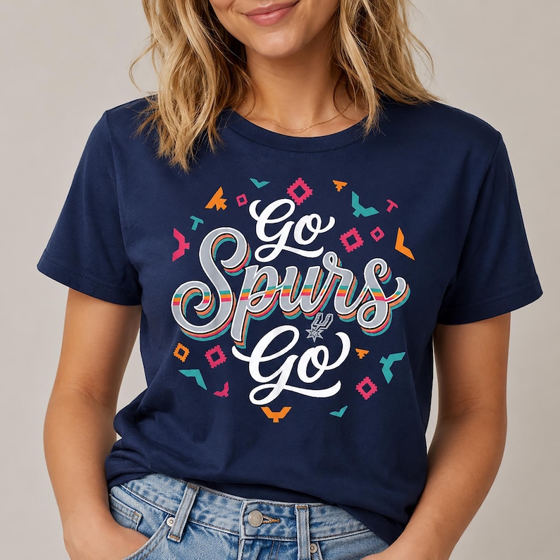 May include: A navy blue t-shirt with the words "Go Spurs Go" in a layered, colourful design. The shirt has a round neck and short sleeves. The design incorporates geometric shapes and the Spurs logo.