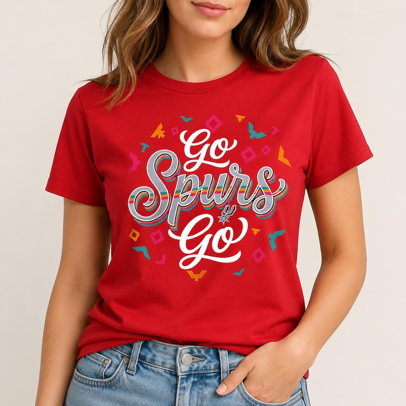 May include: Red t-shirt with the text "Go Spurs Go" in a stylized, multi-coloured design. The shirt features a graphic of the San Antonio Spurs logo and decorative shapes. A casual, short-sleeved top.
