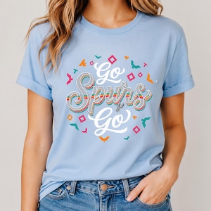 May include: Pale blue t-shirt with rolled sleeves. The shirt displays the text "Go Spurs Go" in a colourful, retro design. The design includes geometric shapes and bird graphics, and has a crew neck.