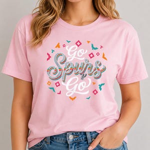 May include: A pale pink t-shirt featuring the text "Go Spurs Go" in a retro style, with a colourful design. The text is encircled by small, geometric shapes in various colours. The San Antonio Spurs logo is included.