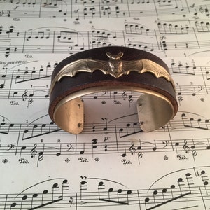 May include: A brown leather cuff bracelet with a silver bat design. The bracelet is on a sheet of music.