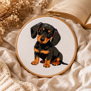 Pode incluir: Um bastidor de bordado circular de madeira contendo um padrão de ponto cruz de um filhote de dachshund preto e castanho. O filhote está sentado com olhos escuros grandes, orelhas caídas e marcas castanhas no rosto, peito e patas.