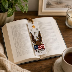 May include: A rectangular fabric bookmark featuring a cross-stitched design of a bald eagle perched atop a red, white, and blue American flag. Above the eagle are four embroidered stars, one large gold star and three smaller silver stars, with a brown tassel.