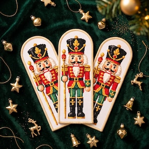 Op de afbeelding: Drie boogvormige borduurornamenten met een notenkraker-soldaat in een rood, groen en zwart uniform met een hoge hoed, gouden accenten en een staf. Elk stuk is afgewerkt met een crèmekleurige gestikte rand op een witte stoffen ondergrond.
