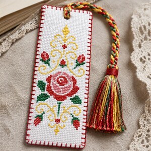 May include: A rectangular white fabric bookmark featuring a cross-stitch design of a central pink rose surrounded by yellow scrollwork and small red rosebuds. The edges are finished with red blanket stitching and a braided cord with a multicoloured tassel.