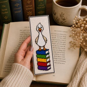 May include: A rectangular cross-stitch bookmark featuring a white duck with orange feet and a yellow beak standing atop a vertical stack of five books in blue, green, yellow, red, and purple, all outlined in black on a white fabric background.
