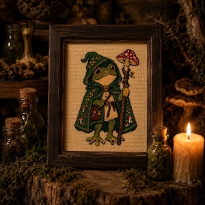 May include: A framed cross-stitch embroidery featuring a green frog wearing a dark green hooded cloak and a beige tunic. The frog holds a wooden staff topped with a red and white spotted mushroom. Small mushrooms are stitched on the cloak.
