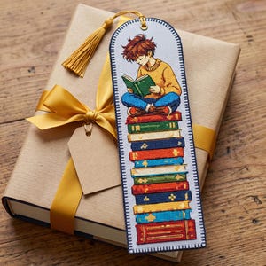 Puede incluir: Un marcapáginas de tela rectangular con una ilustración en punto de cruz de una persona de pelo castaño con un jersey amarillo y pantalones azules, sentada sobre una alta pila de libros coloridos. Tiene una borla dorada en la parte superior.