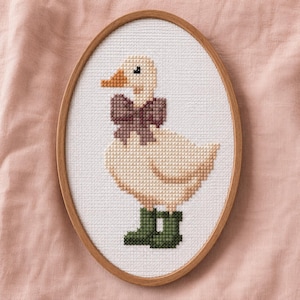 May include: Cross-stitch artwork of a duck wearing a bow and boots, framed in an oval wooden hoop. The duck is cream-colored with an orange beak and black eyes. The bow is a dark mauve color, and the boots are green.