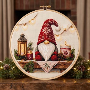 May include: A circular wooden embroidery hoop containing a cross-stitch pattern of a gnome wearing a red patterned hat and mittens. The gnome is centered between a lit lantern, a small candle, and greenery, with string lights in the background.
