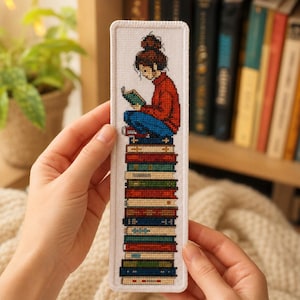 Puede incluir: Un marcapáginas rectangular de tela con un diseño de punto de cruz de una persona con el pelo castaño recogido en un moño, que lleva un suéter rojo y pantalones azules, sentada sobre una alta pila de libros de colores leyendo un libro verde.