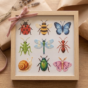 May include: A square light wood frame containing a cross-stitch embroidery on white fabric. The design features nine insects and small creatures: a ladybird, bee, blue butterfly, grasshopper, dragonfly, ant, snail, beetle, and pink moth. A pink ribbon is tied to the top left corner.