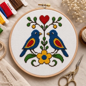 May include: A completed cross-stitch embroidery in a wooden hoop. The design features two blue birds facing each other, a red heart, and a yellow flower. Embroidery floss, scissors, and a needle are visible.