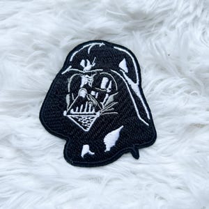 Puede incluir: Parche bordado en blanco y negro con el casco de Darth Vader. El parche presenta detalles intrincados con costuras blancas sobre un fondo negro. Para ropa o accesorios.