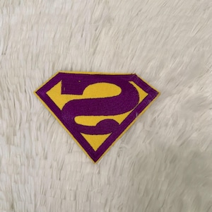 Pode incluir: Um patch bordado amarelo e roxo em forma de diamante. O patch apresenta um fundo amarelo com o número "2" roxo no centro. O patch é provavelmente para roupas ou acessórios.