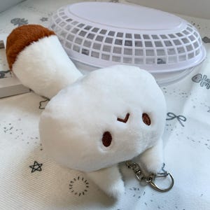May include: A plush keychain shaped like a mushroom with a white cap and a brown top. The mushroom features a simple embroidered face with two brown eyes and a small brown mouth. A silver metal lobster clasp is attached to the bottom of the plush.