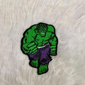 Puede incluir: Parche bordado que representa a Hulk en una pose dinámica. El parche muestra la piel verde de Hulk, pantalones morados y un contorno negro. El diseño captura el físico musculoso y la expresión decidida del personaje, para los fanáticos de los cómics.
