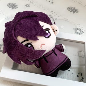Peut inclure: Une poupée en peluche avec des cheveux courts violet foncé et un sweat à capuche violet assorti. La poupée présente des traits du visage brodés, notamment de grands yeux violet clair, des sourcils foncés et une petite bouche sur un visage pâle.