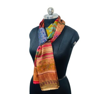 May include: A rectangular scarf featuring a vibrant patchwork of patterns in shades of orange, red, yellow, blue, and green. The design includes horizontal bands of paisley motifs, floral prints, and geometric shapes, draped around a black mannequin.
