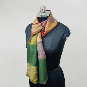 May include: A multi-colored scarf featuring a patchwork design with various floral and geometric patterns in shades of green, tan, red, and purple. The scarf is draped around the neck of a black mannequin, showcasing a mix of printed fabric panels.