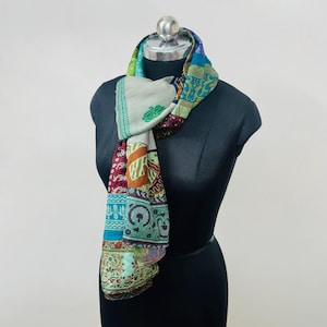 May include: A rectangular scarf featuring a patchwork design with various floral and geometric patterns in shades of teal, cream, orange, and deep red. The scarf is draped around the neck of a black mannequin, displaying a mix of intricate prints.