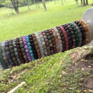 May include: A collection of numerous beaded bracelets featuring a wide variety of natural stone colors and textures, including white, green, blue, purple, brown, and black, all arranged side-by-side on a cylindrical light gray display stand.