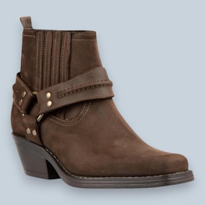 May include: A dark brown ankle boot featuring a square toe, a stacked heel, and a decorative strap across the mid-foot secured with metallic rivets and a side ring. The boot includes vertical elastic side panels and a pull tab at the top rear.