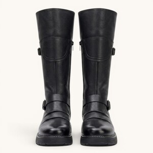 May include: A pair of black knee-high boots. The boots are made of leather and feature a round toe, a chunky sole, and decorative buckles. The boots have a zipper on the side for easy wear.
