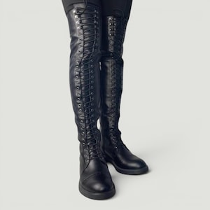 May include: A pair of black over-the-knee boots featuring a full-length front lace-up design with silver-toned metal eyelets. The boots have a rounded toe, a flat sole, and a smooth finish that extends well above the knee with a structured fit.