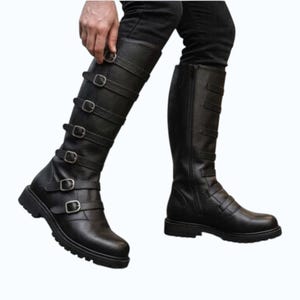 May include: A pair of black knee-high boots featuring a textured leather-like finish, a rounded toe, and a thick lug sole. Each boot is adorned with six horizontal straps across the front, secured by silver-toned metal buckles and side zippers.