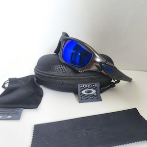 May include: Black and silver sunglasses with blue lenses rest on a black case. Included are a black microfiber cleaning cloth, a black pouch, and a tag with the brand logo. The lenses reflect a bright blue color.