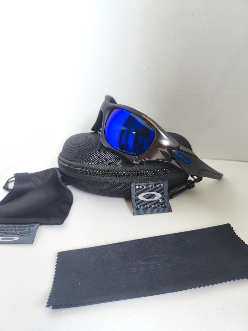 May include: Black and silver sunglasses with blue lenses rest on a black case. Included are a black microfiber cleaning cloth, a black pouch, and a tag with the brand logo. The lenses reflect a bright blue color.
