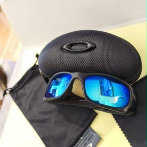 Puede incluir: Gafas de sol negras con lentes azules espejadas, estuche negro, bolsa con cord&oacute;n negra y pa&ntilde;o de limpieza. Dise&ntilde;o deportivo, probablemente para actividades al aire libre. El estuche tiene el logo de la marca.