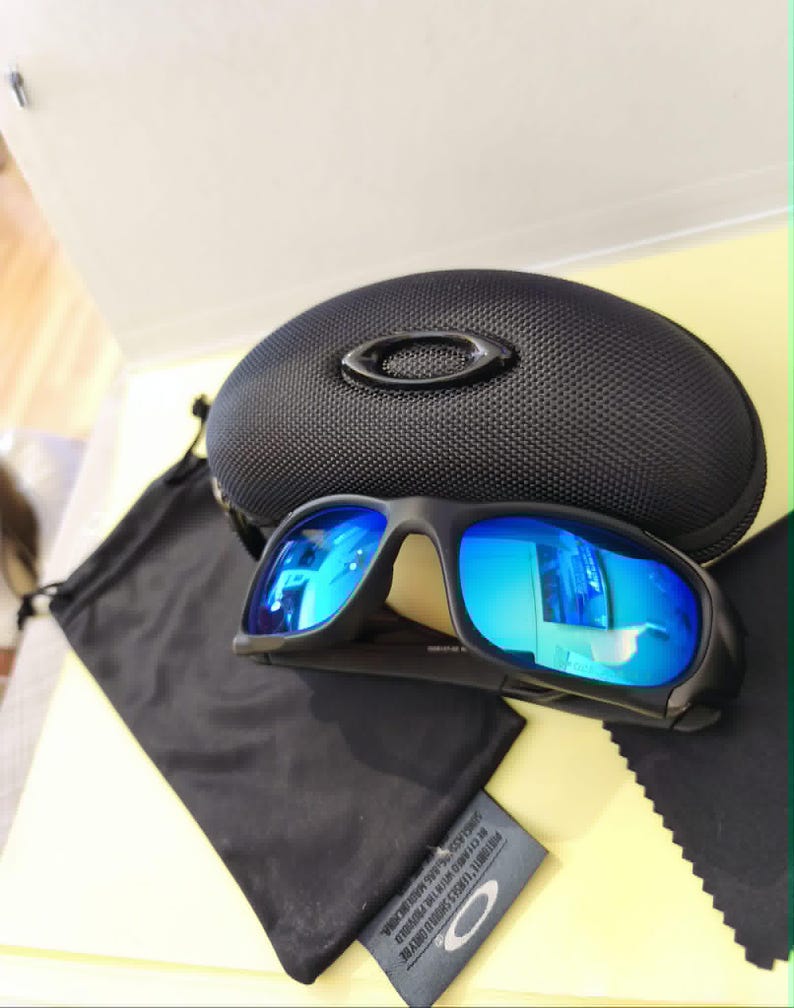 Puede incluir: Gafas de sol negras con lentes azules espejadas, estuche negro, bolsa con cord&oacute;n negra y pa&ntilde;o de limpieza. Dise&ntilde;o deportivo, probablemente para actividades al aire libre. El estuche tiene el logo de la marca.