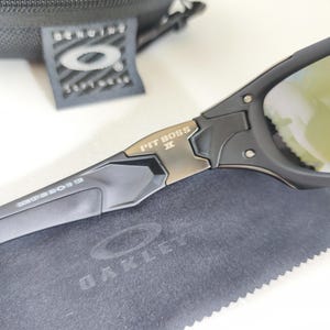 May include: Black Oakley Pit Boss II sunglasses with gold-tinted lenses. The sunglasses have a black frame with the text "PIT BOSS II" in gold. The image also shows a black case, a tag, and a gray cleaning cloth with the Oakley logo.