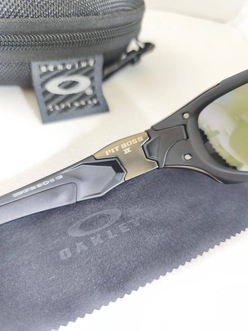 May include: Black Oakley Pit Boss II sunglasses with gold-tinted lenses. The sunglasses have a black frame with the text "PIT BOSS II" in gold. The image also shows a black case, a tag, and a gray cleaning cloth with the Oakley logo.