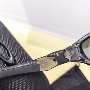 May include: Black sunglasses with the text "PIT BOSS" on the arm. The frame has a sleek, modern design with a silver accent. The lenses are a reflective silver color. The arm of the sunglasses has the numbers "009137-05 60 19 138".