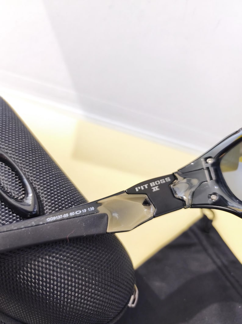 May include: Black sunglasses with the text "PIT BOSS" on the arm. The frame has a sleek, modern design with a silver accent. The lenses are a reflective silver color. The arm of the sunglasses has the numbers "009137-05 60 19 138".