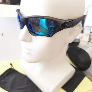 Puede incluir: Gafas de sol negras y grises con lentes reflectantes azules. Las gafas tienen una montura curvada y se muestran en una cabeza de maniqu&iacute; blanca. Una funda negra y un pa&ntilde;o de limpieza son visibles debajo.