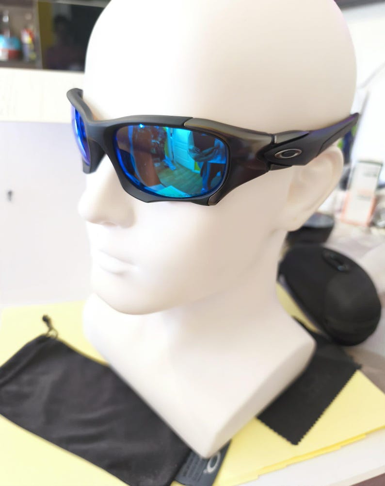 Puede incluir: Gafas de sol negras y grises con lentes reflectantes azules. Las gafas tienen una montura curvada y se muestran en una cabeza de maniqu&iacute; blanca. Una funda negra y un pa&ntilde;o de limpieza son visibles debajo.
