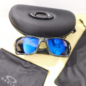 May include: Black sunglasses with blue reflective lenses, a black case, and a black cleaning cloth. The sunglasses have a sporty design. The case has a textured surface and the Oakley logo is visible on the cloth.