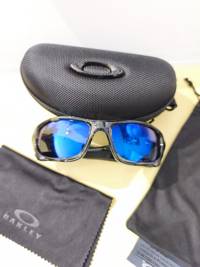 May include: Black sunglasses with blue reflective lenses, a black case, and a black cleaning cloth. The sunglasses have a sporty design. The case has a textured surface and the Oakley logo is visible on the cloth.