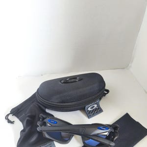 May include: Black sunglasses with blue lenses and accents, a black case, a black drawstring bag, a black cleaning cloth, and a tag. The sunglasses have a sleek, modern design. The case has a zipper closure and the brand logo.