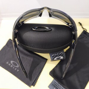 May include: Black sunglasses with a black case, cleaning cloth, and drawstring bag. The sunglasses have a curved design and dark lenses. The case is textured and has a metal emblem. The cleaning cloth has the brand name printed on it.