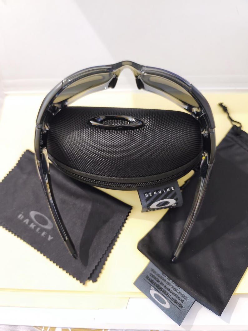 May include: Black sunglasses with a black case, cleaning cloth, and drawstring bag. The sunglasses have a curved design and dark lenses. The case is textured and has a metal emblem. The cleaning cloth has the brand name printed on it.
