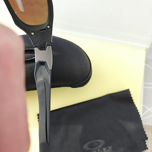 Puede incluir: Gafas de sol negras y grises con lentes de color naranja. Las gafas de sol son parcialmente visibles, junto con un estuche negro y un pa&ntilde;o de limpieza negro con el logotipo de Oakley.