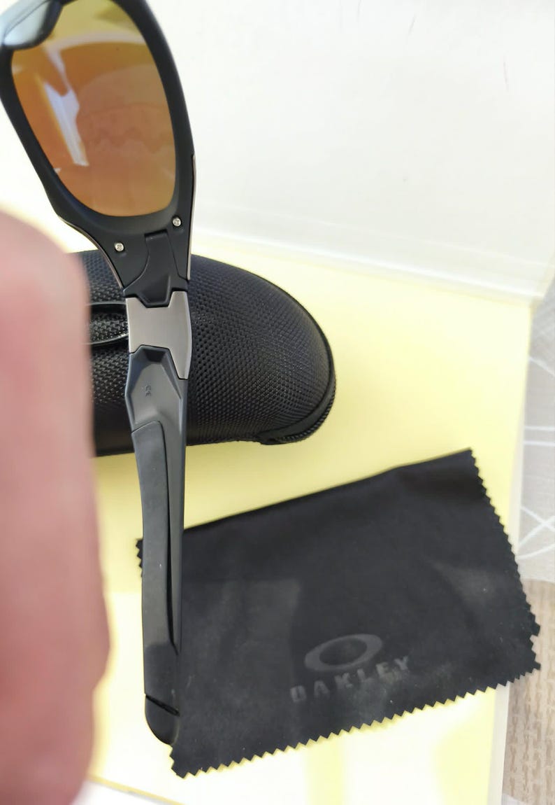 Puede incluir: Gafas de sol negras y grises con lentes de color naranja. Las gafas de sol son parcialmente visibles, junto con un estuche negro y un pa&ntilde;o de limpieza negro con el logotipo de Oakley.