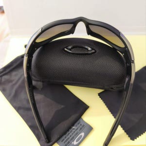 Puede incluir: Gafas de sol negras con montura curvada y lentes degradadas, sobre un estuche negro. Se incluye un pa&ntilde;o de limpieza de microfibra negro y una bolsa con cord&oacute;n negra. El logotipo de la marca es visible.
