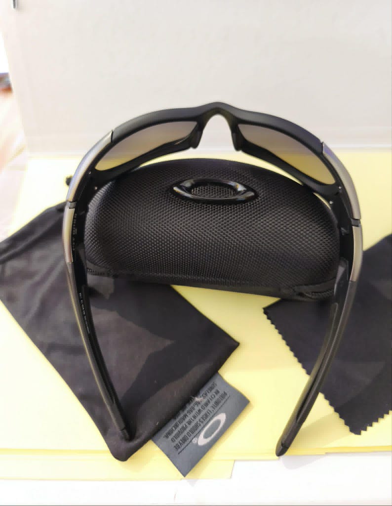 Puede incluir: Gafas de sol negras con montura curvada y lentes degradadas, sobre un estuche negro. Se incluye un pa&ntilde;o de limpieza de microfibra negro y una bolsa con cord&oacute;n negra. El logotipo de la marca es visible.
