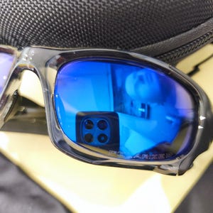 May include: Close-up of polarized sunglasses with blue lenses and a black frame. The lenses reflect a bright blue hue, and the text "POLARIZED" is visible on the lens. The sunglasses are resting on a black case.